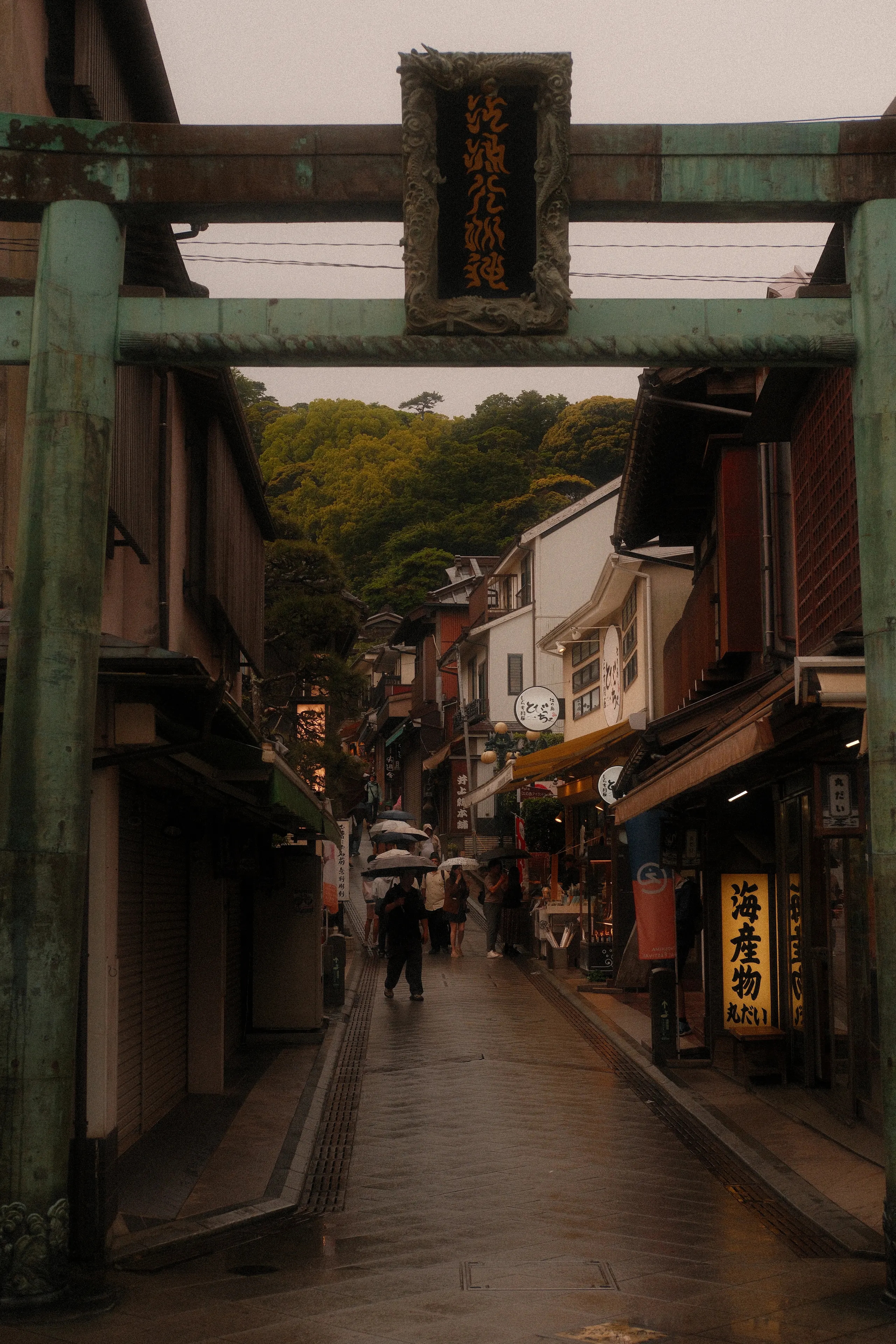 A weathered bronze torii over Enoshima's rainy main street